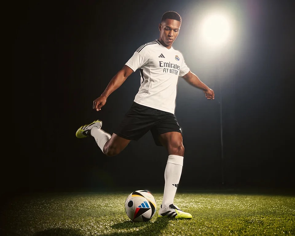 Male soccer player in a white Adidas jersey, black shorts, and yellow cleats, kicking a colorful soccer ball.