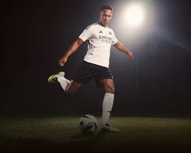 Male soccer player in a white Adidas jersey, black shorts, and yellow cleats, kicking a colorful soccer ball.